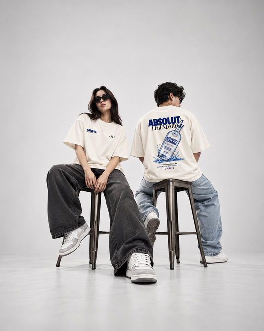 Two models sitting on stools in a studio setting, wearing oversized off-white graphic t-shirts. The model on the left wears a small chest graphic and dark gray wide-leg jeans, while the model on the right wears a large back graphic of Absolut Transfers and light blue jeans.