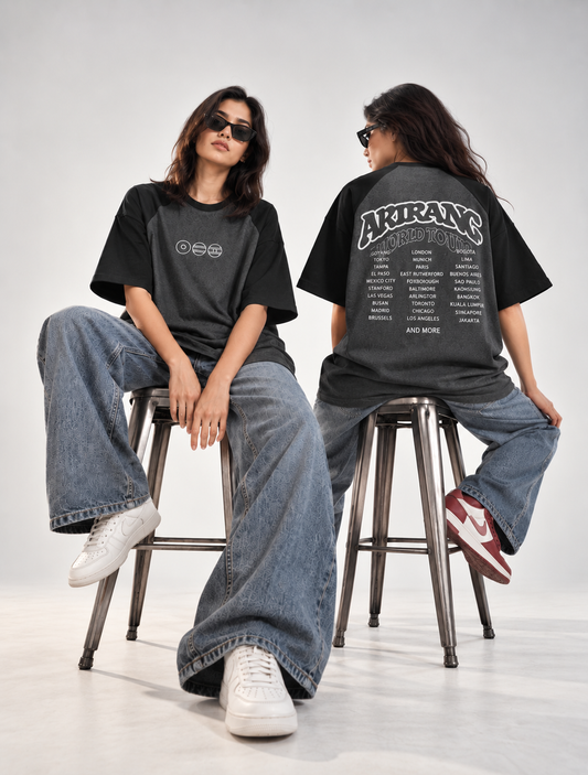 Two models wearing black oversized graphic t-shirts and wide-leg blue jeans, seated on metal stools in a studio setting. One model shows the front of the shirt with a small white logo, and the other shows the back with a large white graphic text reading 'AKIRANS COPENHAGEN'.