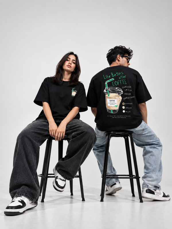 A man and a woman wearing black graphic t-shirts and jeans, seated on black metal bar stools in a studio setting. The woman wears a black t-shirt with a small green and white chest graphic, while the man wears a black t-shirt with a large back graphic depicting a coffee cup and the text 'Life begins after COFFEE'.