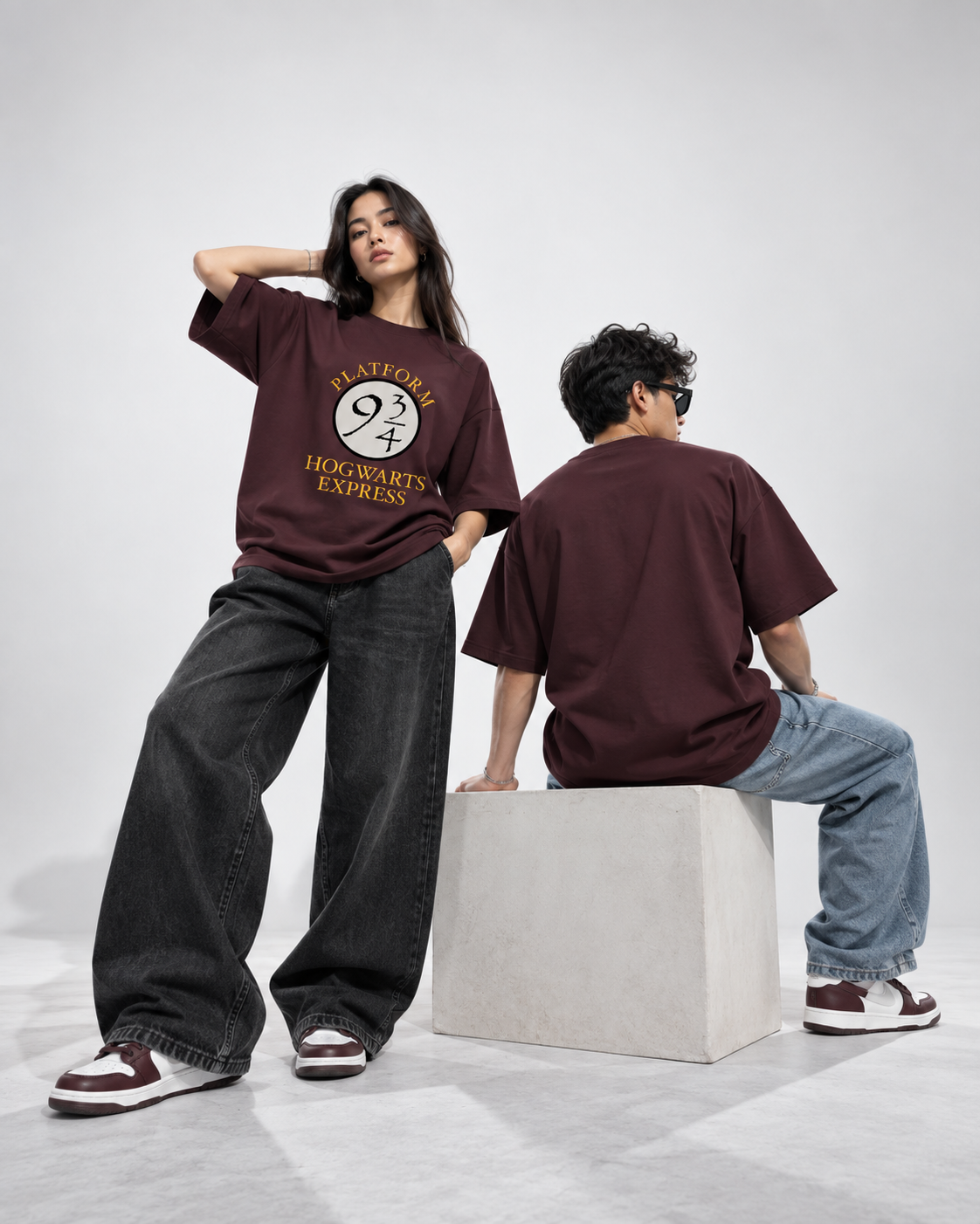Two models wearing oversized maroon graphic t-shirts and wide-leg jeans pose in a minimalist studio setting. One model stands facing forward, showcasing the front graphic of the shirt which reads 'PLATFORM 97 EXPRESS' in yellow and white.