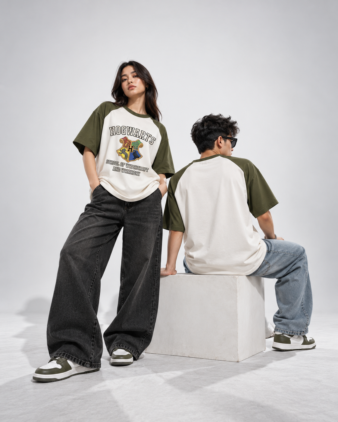 Two models wearing a raglan-style t-shirt with olive green sleeves and a cream body featuring a graphic print of a cartoon bear and the text 'HONESTHEART'. One model stands facing forward, and the other sits on a white block facing away, in a studio setting.