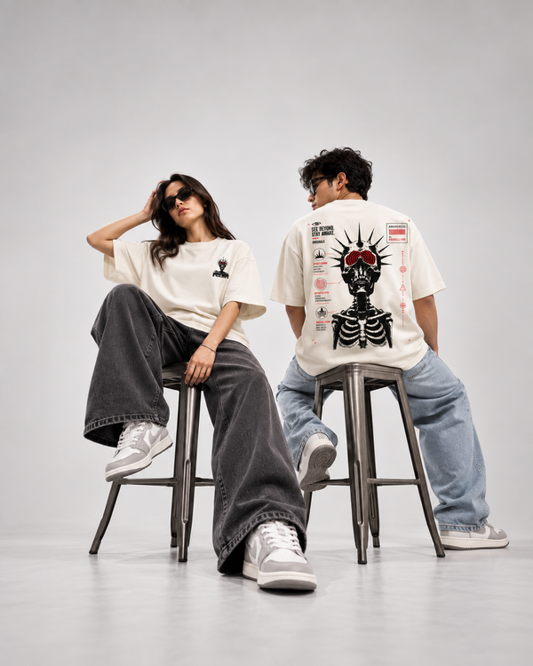 A man and a woman sit on metal stools in a studio setting, wearing oversized white graphic T-shirts and jeans. The woman wears a black-and-white graphic tee with dark gray wide-leg jeans and white sneakers.