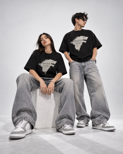 A man and a woman modeling matching black graphic t-shirts with a white direwolf logo and 'WINTER IS COMING' text, styled with wide-leg gray jeans and gray sneakers against a light gray studio background.
