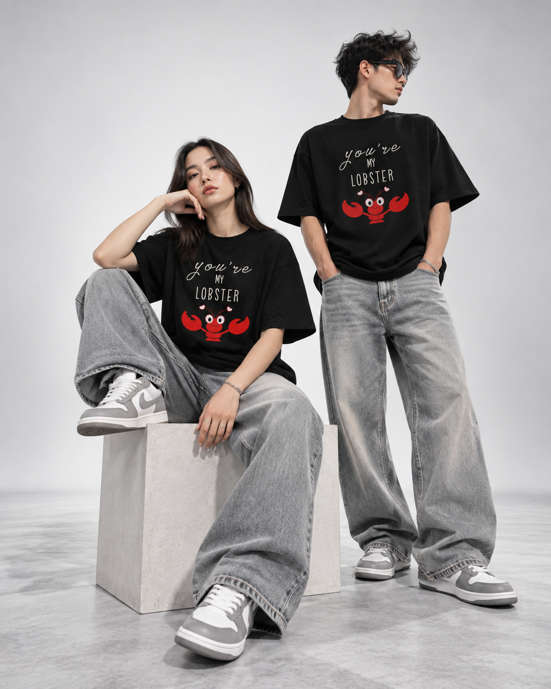 Two models wearing matching black graphic t-shirts with a red lobster design and the text 'You're My LOBBYER', styled with wide-leg gray jeans and sneakers in a studio setting.
