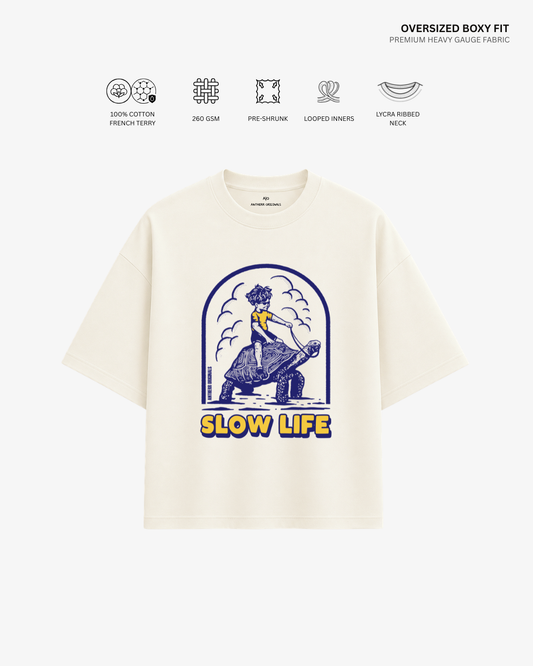 An oversized short-sleeve T-shirt in cream-colored heavy cotton fabric, featuring a blue graphic of a person riding a horse with the text 'SLOW LIFE' in yellow. The image includes technical feature icons and text at the top against a clean white background.